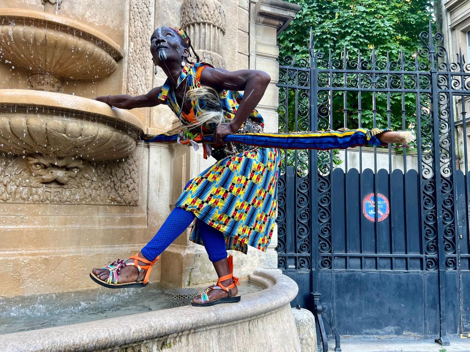 Kemet en tenue wax, le bâton tendu à l'horizontale devant une fontaine parisienne